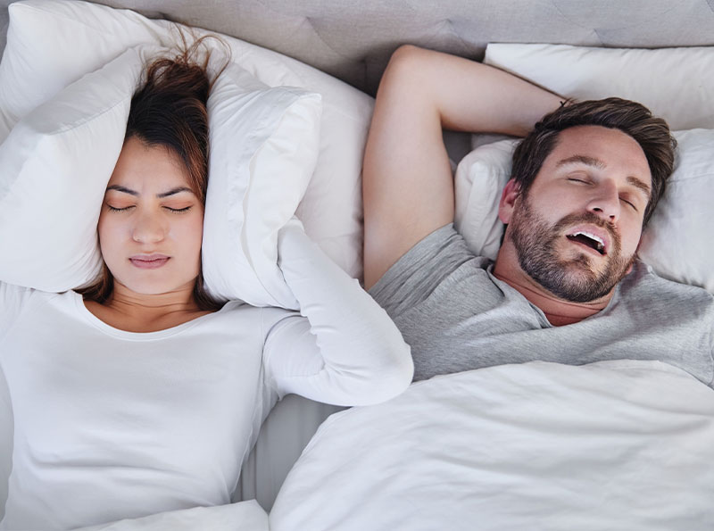 Man snoring next to woman covering her ears with a pillow